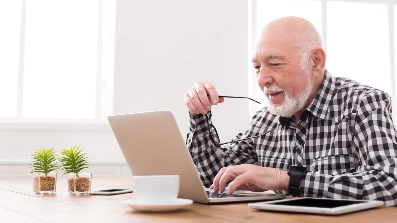 Ein Seniorin sitzt am Esstisch und schaut in seinen Computer